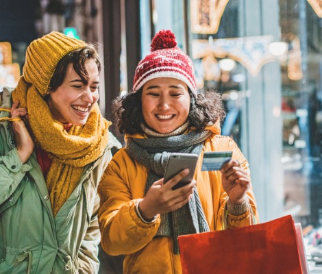 Two women holiday shopping outside using credit card and smart phone