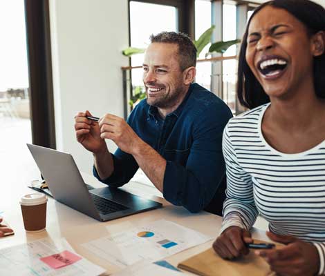 two coworkers laugh while drinking coffee and looking at laptops